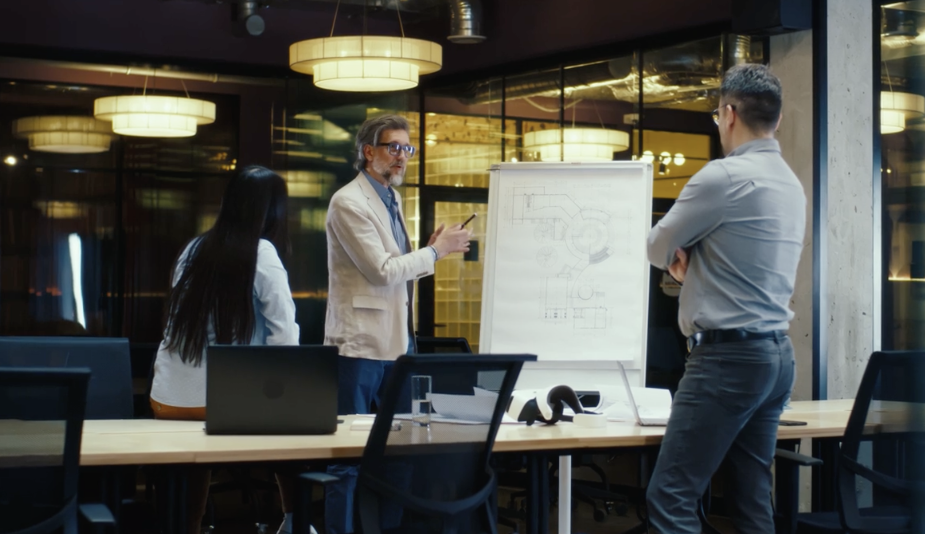 Three colleagues discussing diagrams on a presentation board in a glass-walled office